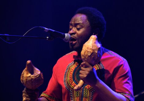 A person in a vibrant traditional outfit sings into a microphone while playing a pair of gourd maracas on stage.