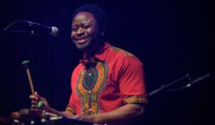 A man wearing a red patterned shirt plays a percussion instrument while smiling into a microphone on stage.