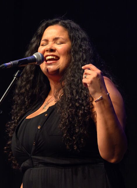 A woman with long curly hair sings passionately into a microphone on stage, wearing a black sleeveless top.