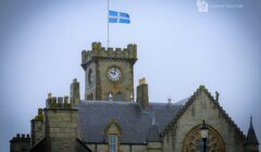 A stone building with a clock tower, featuring a blue and white flag at the top. Text in the image reads "Lerwick Town Hall.