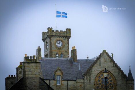 A stone building with a clock tower, featuring a blue and white flag at the top. Text in the image reads "Lerwick Town Hall.