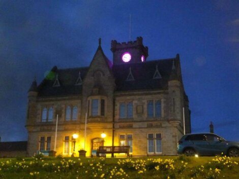 A stone building with a lit clock tower stands against a dark blue evening sky. Warm lights illuminate the entrance, and a car is parked to the right. Yellow flowers are visible in the foreground.