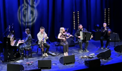 Five musicians perform on stage under blue lighting. They play various instruments including a piano, a flute, a violin, an accordion, and a double bass.