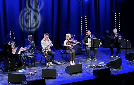 Five musicians perform on stage under blue lighting. They play various instruments including a piano, a flute, a violin, an accordion, and a double bass.