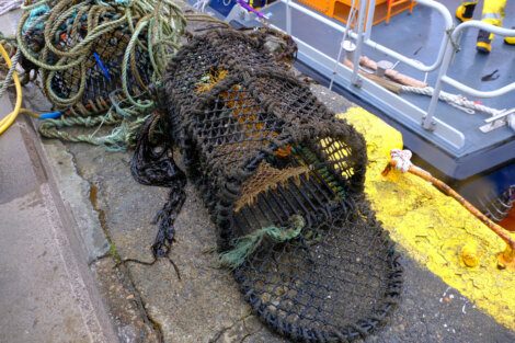 A worn fishing net trap is placed on a concrete dock near a yellow-painted edge, with tangled ropes and a nearby boat visible in the background.