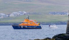 An orange and blue lifeboat assists a small white boat near a rocky coastline with buildings and green hills in the background.