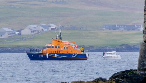 An orange and blue lifeboat assists a small white boat near a rocky coastline with buildings and green hills in the background.