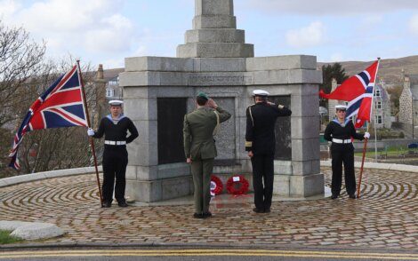 Four individuals, including two holding British and Norwegian flags, salute a stone monument with wreaths at its base. The ceremony takes place on a cobblestone platform in a town setting.