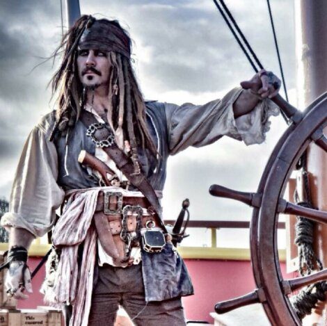 A person dressed in a pirate costume, complete with a hat, dark eye makeup, and various belts and accessories, stands at the helm of a ship, holding the wheel.