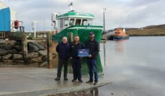 Three men standing on a pier holding a sign. A green boat and an orange boat are docked on the water in the background. The sky is cloudy.