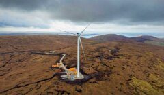 An aerial view of a single wind turbine situated in a vast, barren landscape with cloudy skies overhead. Construction equipment is visible at the base of the turbine.