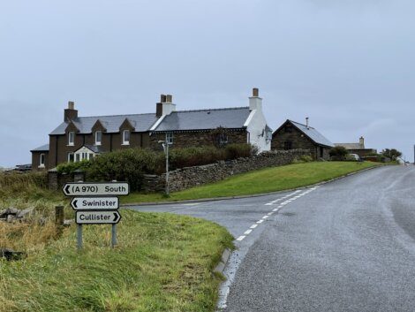 A road with direction signs for South (A970), Swinister, and Cullister near houses on a grassy landscape under an overcast sky.