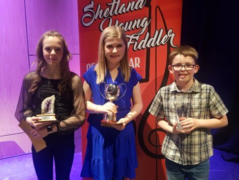 Three young individuals stand holding trophies in front of a "Shetland Young Fiddler of the Year" sign. The boy on the right wears glasses, and the girl in the middle holds a cup trophy.