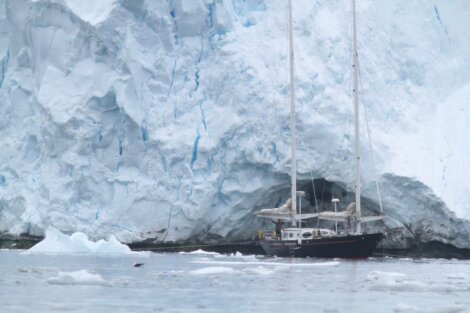 A sailboat is anchored near a massive glacier, surrounded by icy waters and floating ice chunks. The glacier's wall looms in the background.