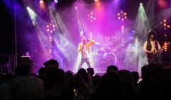 A band performs on a stage with colorful lights and fog effects. The singer at the center wears a yellow jacket, and the guitarist on the right wears a white shirt and long curly hair wig.