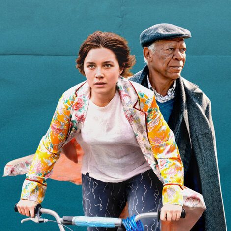 A woman in a floral jacket rides a bicycle with a man in a dark coat and hat sitting behind her.