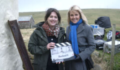 Two women stand outdoors on a film set. One woman holds a clapperboard labeled "Shetland 8." Equipment and a rural landscape are visible in the background.