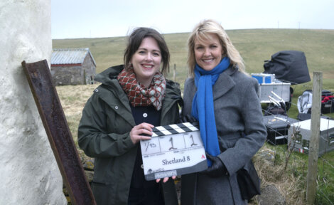 Two women stand outdoors on a film set. One woman holds a clapperboard labeled "Shetland 8." Equipment and a rural landscape are visible in the background.