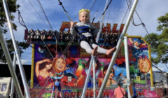 A child, strapped in a harness, is jumping on a bungee ride at a carnival with a vibrant amusement attraction in the background.