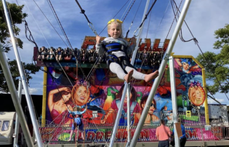 A child, strapped in a harness, is jumping on a bungee ride at a carnival with a vibrant amusement attraction in the background.