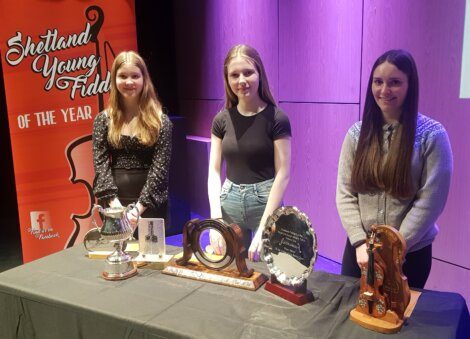 Three young women stand behind a table displaying multiple awards, with a "Shetland Young Fiddler of the Year" banner in the background.