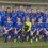 A youth girls' soccer team poses in blue uniforms for a group photo on the field in front of a building labeled "Away Team.