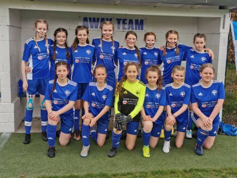 A youth girls' soccer team poses in blue uniforms for a group photo on the field in front of a building labeled "Away Team.