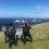 Three people in outdoor clothing stand on a grassy hill overlooking the ocean and rocky islands, with one reading an informational sign.