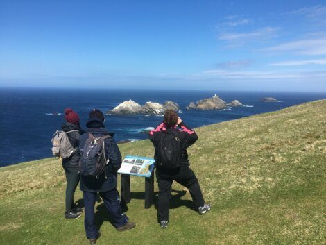 Three people in outdoor clothing stand on a grassy hill overlooking the ocean and rocky islands, with one reading an informational sign.