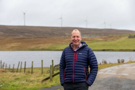 A man in a blue puffer jacket stands on a rural pathway with a lake and wind turbines in the background.