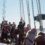 A group of people on a boat work together to raise a sail, with some observing and others taking photos. They are all wearing jackets and life vests. The sky is clear and the sea is visible in the background.