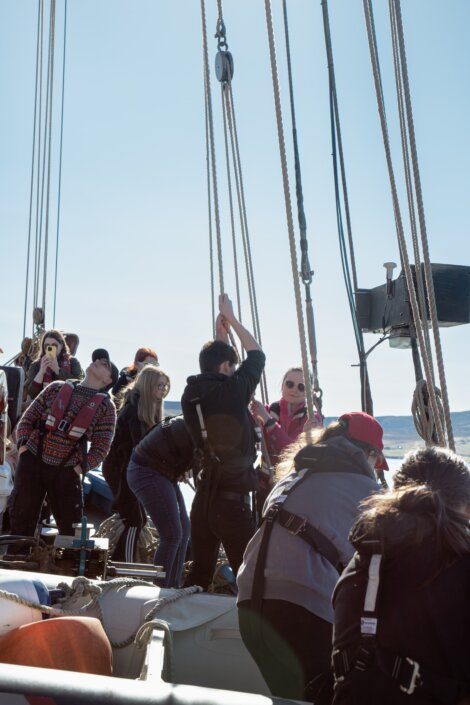 A group of people on a boat work together to raise a sail, with some observing and others taking photos. They are all wearing jackets and life vests. The sky is clear and the sea is visible in the background.