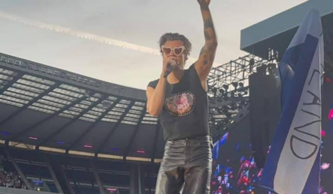 Person in a sleeveless shirt and sunglasses singing on a large stadium stage while raising one hand.