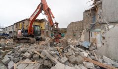 A construction excavator demolishes a partially destroyed building, with debris scattered across the site.