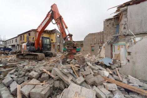 A construction excavator demolishes a partially destroyed building, with debris scattered across the site.