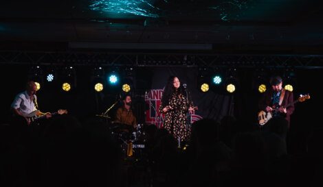 A four-member band performs on stage with instruments and a female vocalist under dim lighting and spotlights.