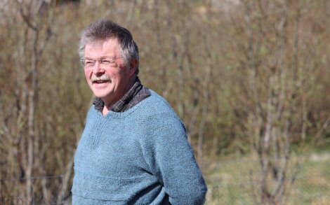 A man with gray hair and glasses wearing a blue sweater stands outside, with blurred trees and foliage in the background.