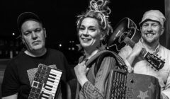 Black and white photo of three people posing with musical instruments: one holds a small keyboard, another smiles with an accordion, and the third carries an acoustic guitar, all facing the camera.