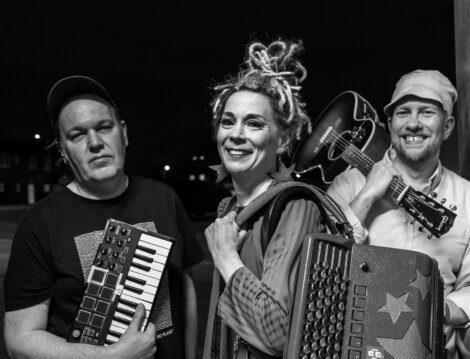 Black and white photo of three people posing with musical instruments: one holds a small keyboard, another smiles with an accordion, and the third carries an acoustic guitar, all facing the camera.