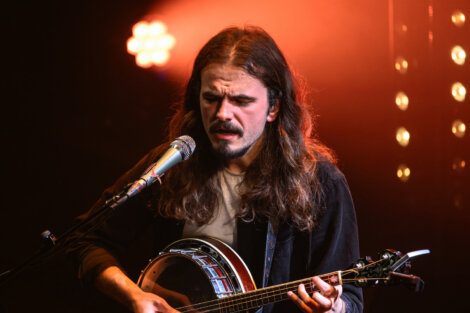 A man with long hair plays a banjo and sings into a microphone under stage lighting.