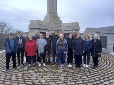 A group of 15 people stands together on a cobblestone platform in front of a large stone monument. It appears to be a cloudy day, and they are all wearing casual clothing and jackets.