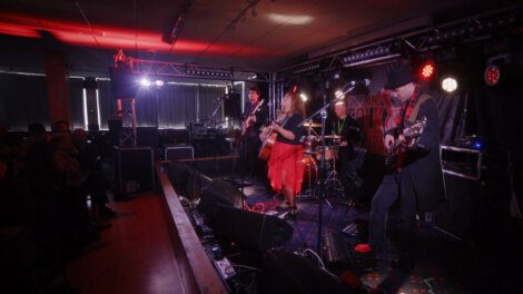 A band performs on a dimly lit stage with guitars and drums, highlighted by red and purple lighting, in front of an audience.