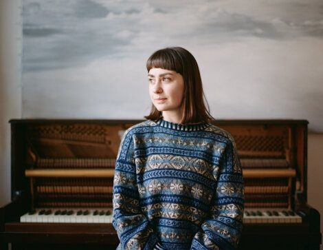 A woman with short hair and a patterned sweater sits in front of an upright piano with its front cover removed, exposing the strings and hammers.