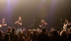 A band performs on stage with a guitarist, a percussionist, a violinist, and a bass player in front of an audience at a concert. Stage lights illuminate the scene.