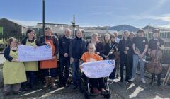 A group of people stand outdoors holding two large checks. One check is for £4,060 to COPE, and the other is for £5,100 to MS Society Scotland. Some members hold musical instruments.