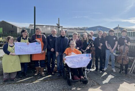 A group of people stand outdoors holding two large checks. One check is for £4,060 to COPE, and the other is for £5,100 to MS Society Scotland. Some members hold musical instruments.