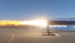 A rocket engine test is underway, emitting a bright plume of flame and exhaust on a clear day, with a shipping container in the background.