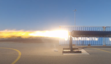 A rocket engine test is underway, emitting a bright plume of flame and exhaust on a clear day, with a shipping container in the background.