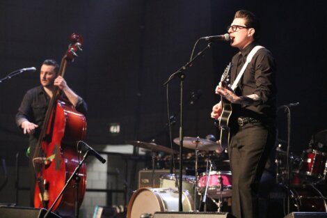 Musician playing an electric guitar and singing into a microphone on stage, accompanied by another musician playing a double bass. Drums and other musical equipment are visible in the background.