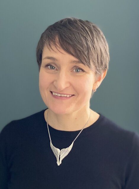 A person with short brown hair smiling and wearing a black shirt and silver wing-shaped necklace stands in front of a blue background.